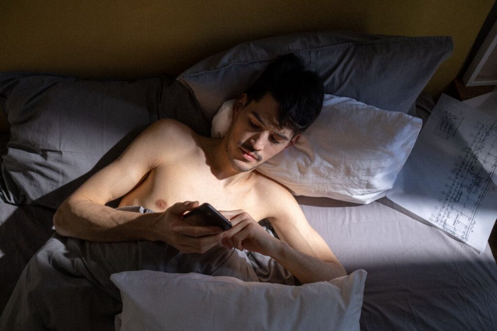 a young man scrolls on phone in bed with sliver of morning light crossing his upper body.